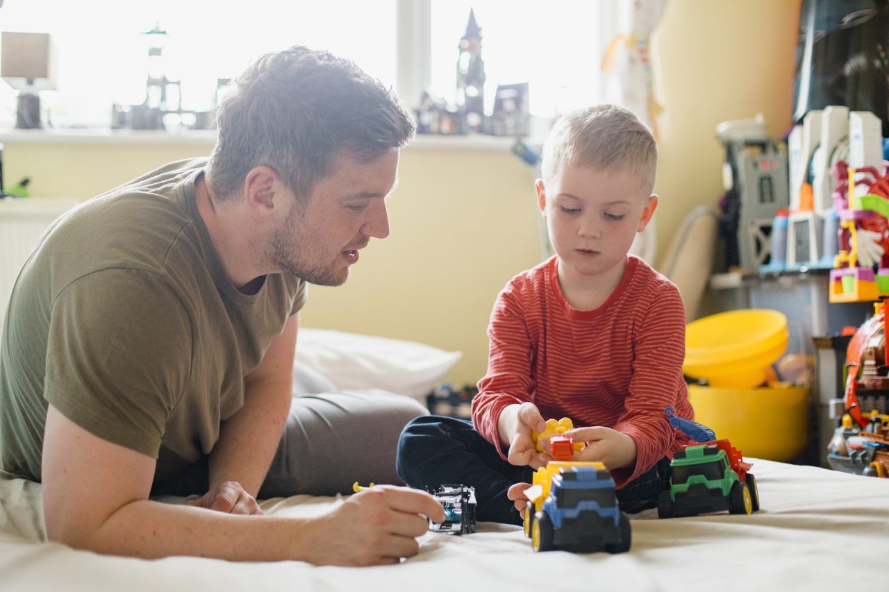 Little boy and his father sitting on his bed in the bedroom and playing with some toy trucks.